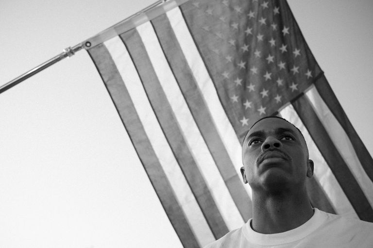 Black and white photo of rapper Vince Staples looking up in a white t-shirt with an American flag behind him, photographed by Adrian Nieto.
